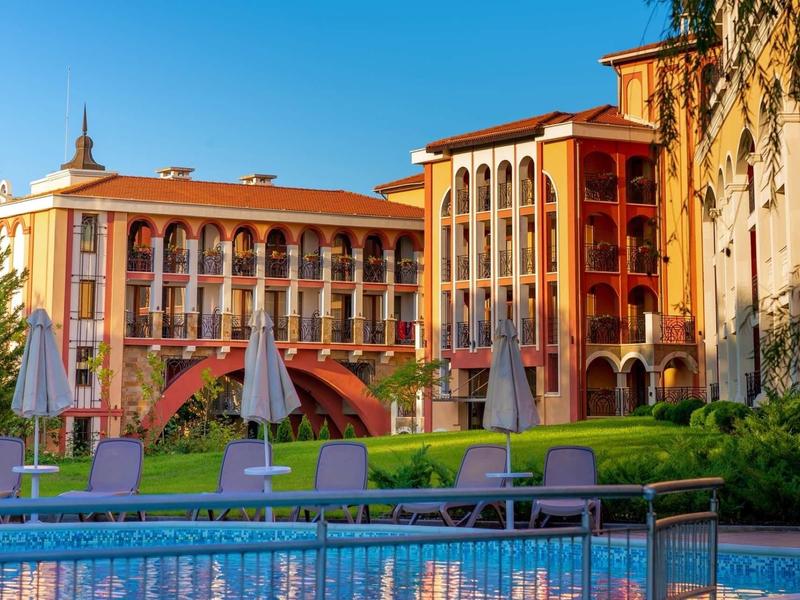 Hotelgebäude mit bogenförmigen Fenstern und Terrasse, davor Pool mit Liegestühlen und Sonnenschirmen.