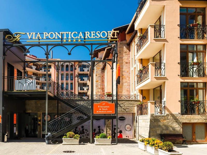 Eingang zum Via Pontica Resort mit Treppen, Balkonen, Blumen und orangefarbenem Hinweisschild.