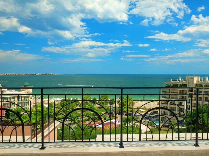 Blick auf blauen Himmel, Meer und grüne Gartenanlage hinter schwarzem Balkon mit Rundbogenmuster.