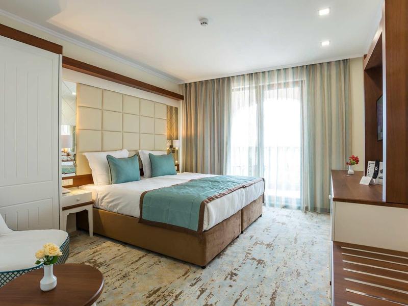 Helles Hotelzimmer mit Doppelbett, Vorhängen, Teppichboden, Tisch und Stuhl, moderne Einrichtung.