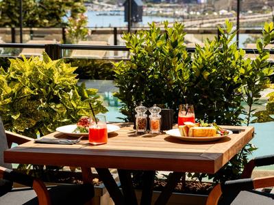 Ein sonniger Terrassentisch mit Getränken und Snacks neben einem blauen Pool und grünen Pflanzen.