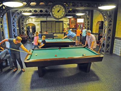 Eine Gruppe von Menschen spielt und beobachtet Billard in einem gemütlichen Raum mit Uhr und Ziegelwänden.