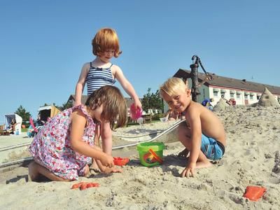 Drei Kinder spielen am sonnigen Strand im Sand mit Eimern und Förmchen.