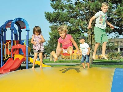 Vier Kinder springen auf einem großen, bunten Hüpfkissen im Freien neben einem Spielplatz.