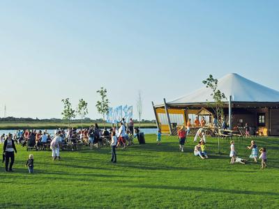 Menschen spielen und entspannen auf einer großen grünen Wiese vor einem modernen Pavillon bei klarem Himmel.