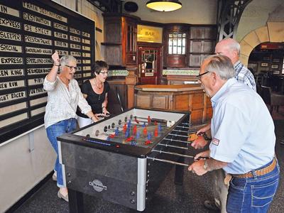 Zwei Paare spielen Tischfußball in einer Bar mit Holzvertäfelung und Getränkekarte an der Wand.