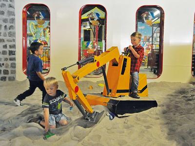 Drei Kinder spielen in einer Sandgrube mit einem gelben Spielbagger vor bunten spiegelnden Fenstern.