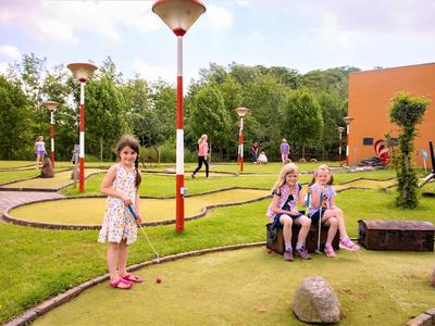 Kinder spielen Minigolf im Freien auf einem grünen Platz mit Bäumen und Gebäuden im Hintergrund.