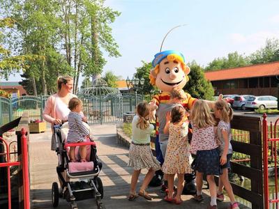 Kinder umgeben eine lebensgroße, fröhliche Maskottchenfigur im Freien bei sonnigem Wetter.