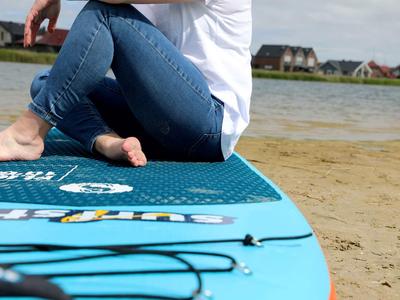 Person sitzt barfuß auf einem blauen SUP-Board am Sandstrand neben Wasser.
