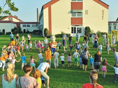 Viele Kinder und Erwachsene spielen und stehen auf einer grünen Wiese vor einem großen Gebäude.