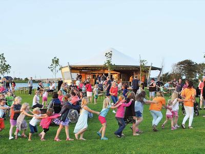 Viele Kinder spielen ein Fangspiel auf einer grünen Wiese vor einem Holzhäuschen.