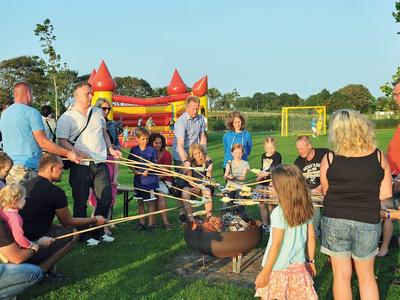 Gruppe Kinder und Erwachsene rösten Stockbrot an offenem Feuer auf grünem Feld bei Sonnenschein.