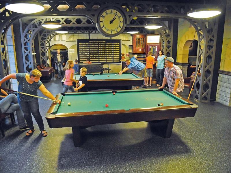 Mehrere Personen spielen in einem Raum mit mehreren Billardtischen unter großen Lampen.