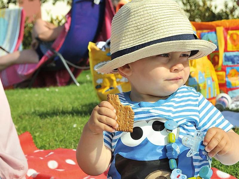 Ein Kleinkind sitzt auf roter Decke im Gras, trägt gestreiftes Shirt, Sonnenhut und hält ein gelbes Spielzeug.