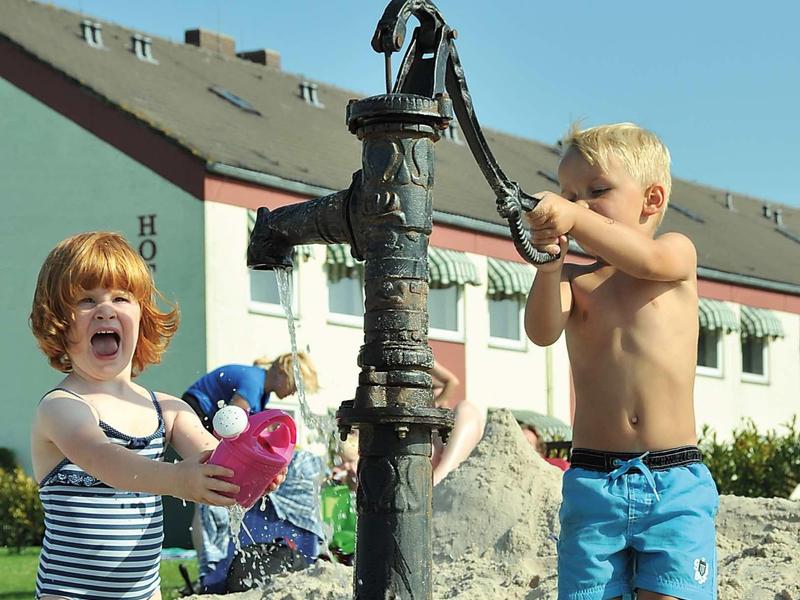 Kinder spielen mit einem alten Wasserpumpenbrunnen am Sandstrand bei klarem Himmel.