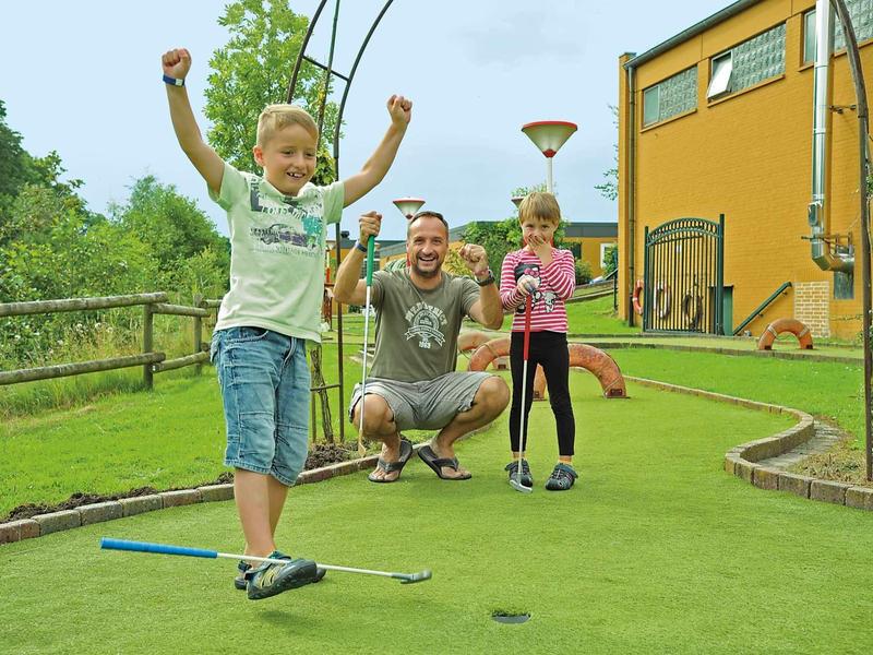 Drei Kinder spielen Minigolf auf grünem Kunstrasen vor einem gelben Gebäude mit hohem Zaun.