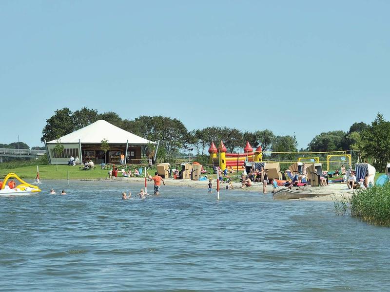 Badende und spielende Menschen an einem Seeufer mit Zelten und Grünfläche im Hintergrund.