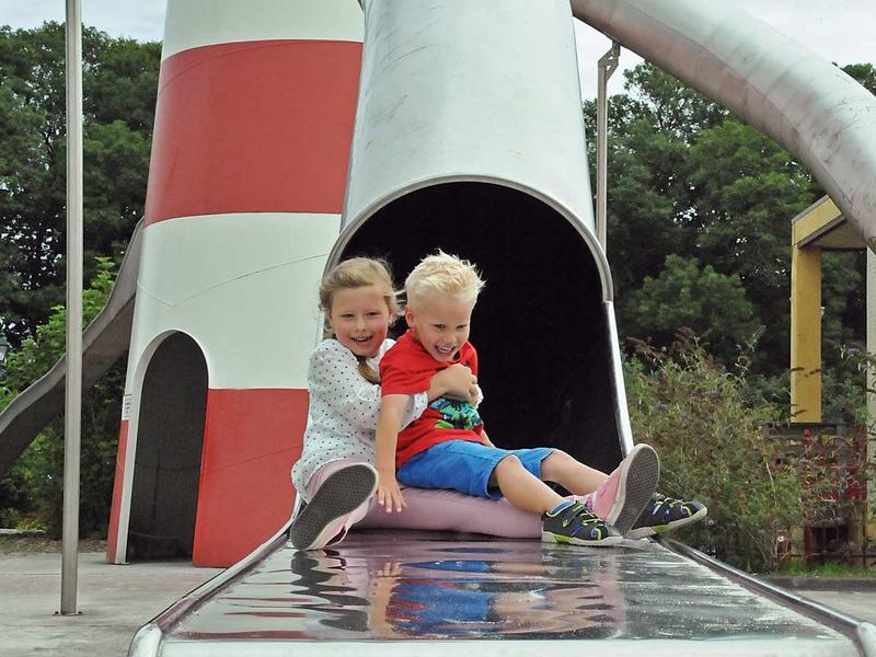 Zwei Kinder sitzen lachend auf einem grauen Metallrutsche-Ausgang an einem Leuchtturmspielplatz.