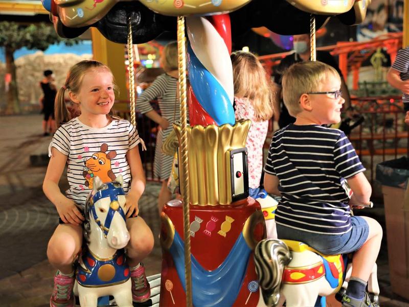 Kinder lachen auf bunten Karussellpferden unter warmem Licht in einem Freizeitpark.
