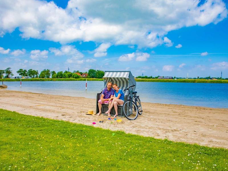Zwei Personen sitzen in einem Strandkorb am Sandstrand neben einem See unter blauem Himmel.