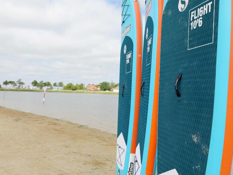 Drei blaue Stand-Up-Paddle-Boards mit orangen Rändern stehen am Ufer eines ruhigen Sees.
