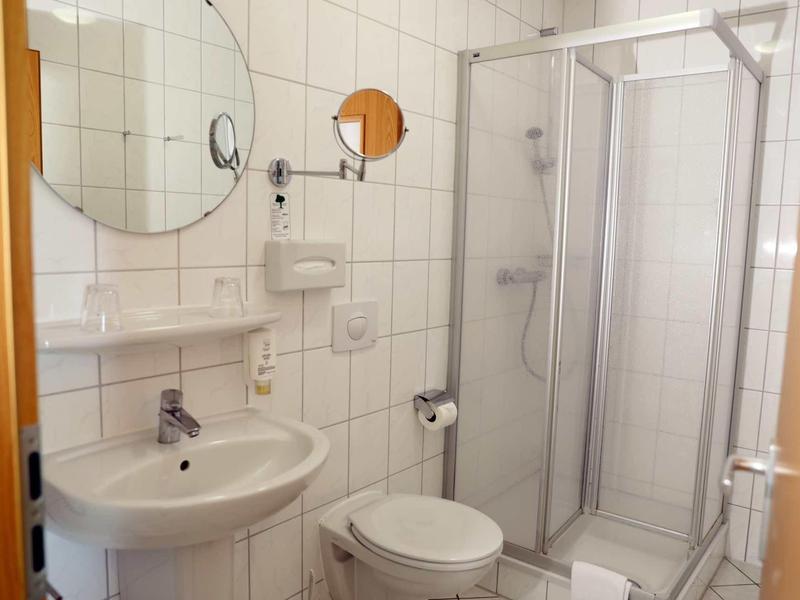 Kleines weiß gekacheltes Badezimmer mit Dusche, Waschbecken, Toilette und rundem Spiegel.