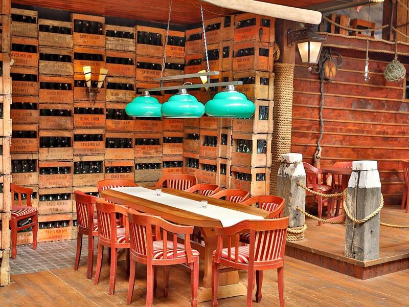 Rustikaler Raum mit Holzwänden, Weinkistenregal, großem Holztisch, roten Stühlen und grünen Hängelampen.