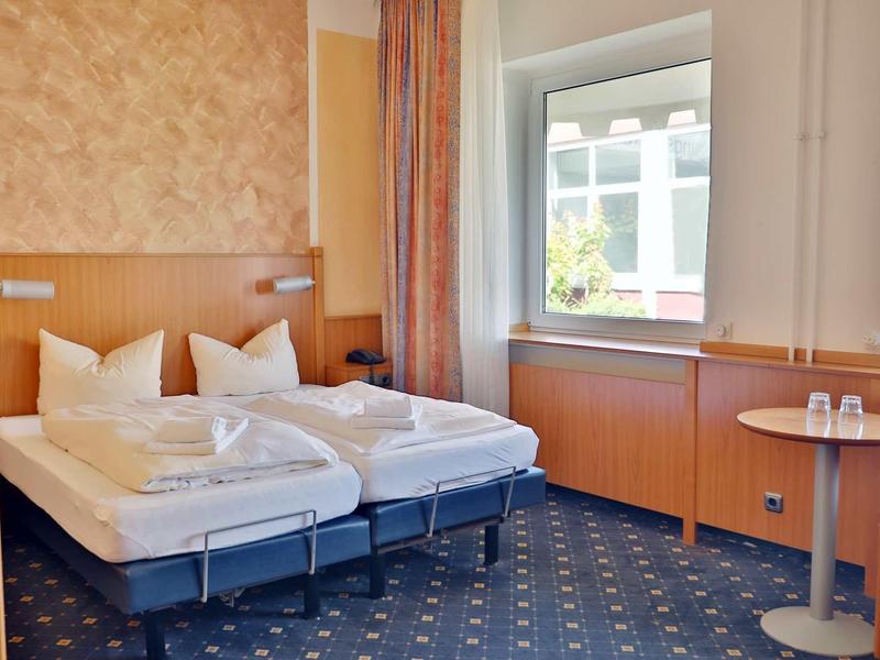 Helles Hotelzimmer mit zwei Einzelbetten, blauem Teppich und Fenster mit Blick ins Freie.