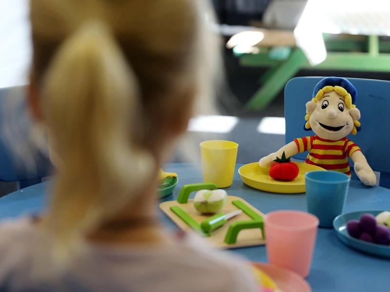 Kind mit Zopf sitzt am Tisch, vor ihm bunte Teller mit Obst und eine Handpuppe.