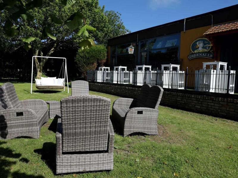 Garten mit grauen Geflechtstühlen und einer weißen Schaukelbank auf grünem Gras vor einem Gebäude.