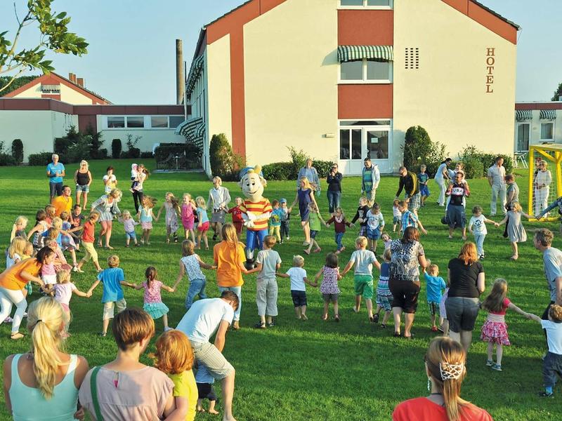 Viele Kinder und Erwachsene spielen und stehen auf einer grünen Wiese vor einem großen Gebäude.