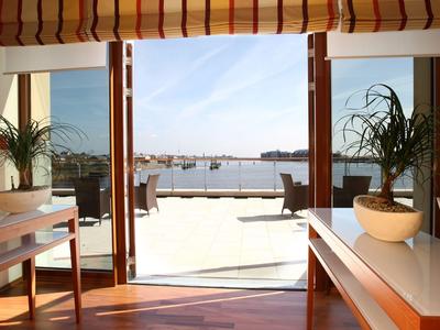 Heller Raum mit großen Fenstertüren, die auf eine sonnige Terrasse mit Blick auf das Wasser führen.