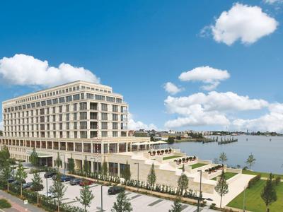 Modernes, großes Gebäude am Ufer mit blauem Himmel, Grünflächen und Wasser im Hintergrund.