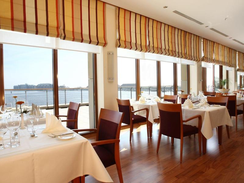 Elegantes Restaurant mit großen Fenstern, Holzboden, weißen Tischen und roten Stühlen am Wasser.