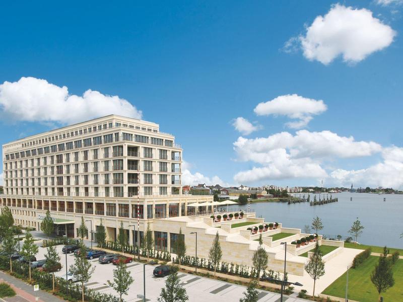 Modernes, großes Gebäude am Ufer mit blauem Himmel, Grünflächen und Wasser im Hintergrund.