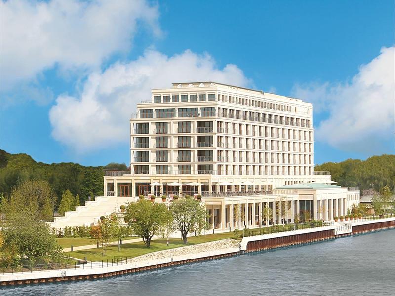 Großes weißes Gebäude am Wasser mit vielen Fenstern, umgeben von Bäumen und blauem Himmel.