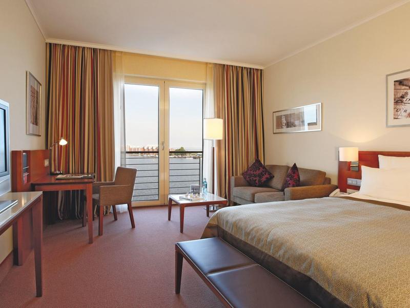 Gemütliches Hotelzimmer mit Doppelbett, Sofa, Schreibtisch, Fernseher und Balkon bei Tageslicht.