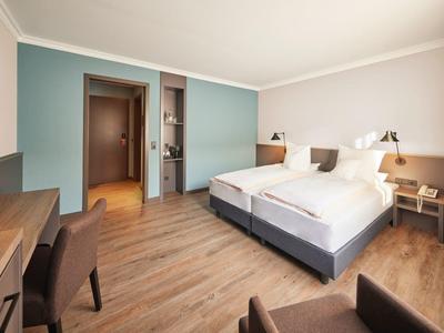 Camera d'hotel luminosa con due letti singoli, pavimento in legno, scrivania e parete d'accento azzurro chiaro