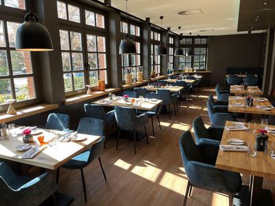 Interno luminoso di un ristorante con tavoli in legno, sedie blu e grandi finestre che lasciano entrare la luce.