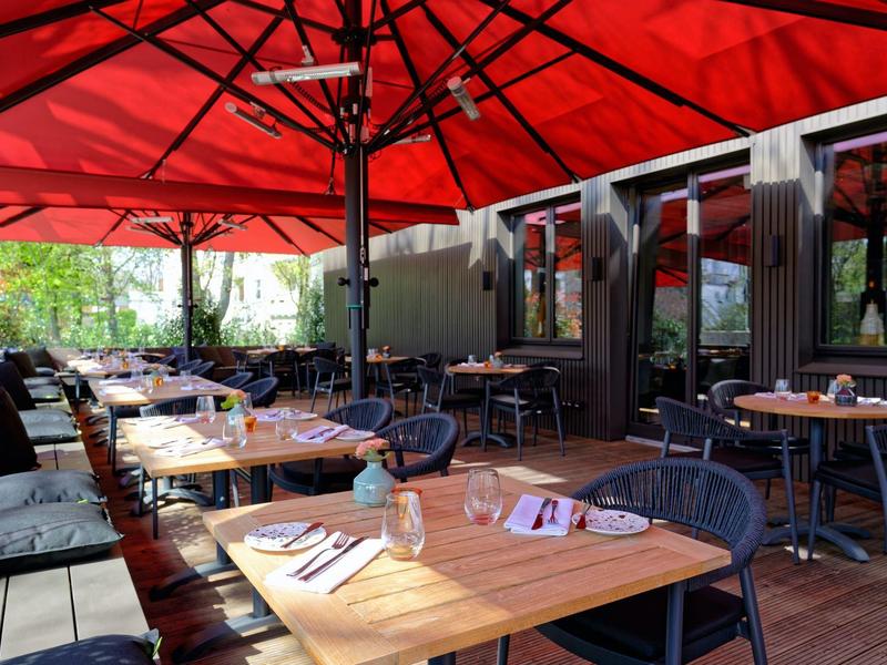 Area pranzo all'aperto con tavoli di legno, sedie e grandi ombrelli rossi su un patio.