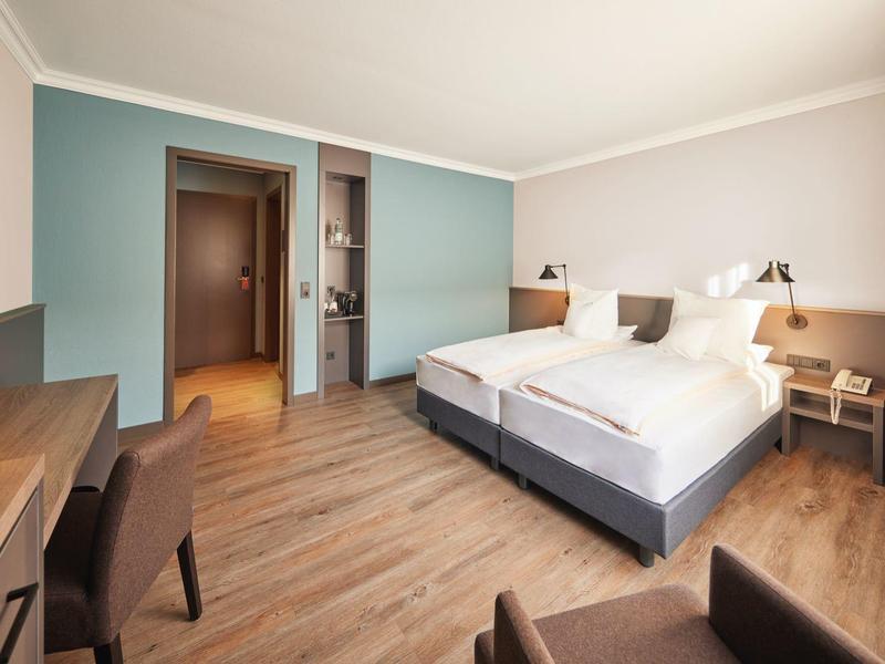 Camera d'hotel luminosa con due letti singoli, pavimento in legno, scrivania e parete d'accento azzurro chiaro