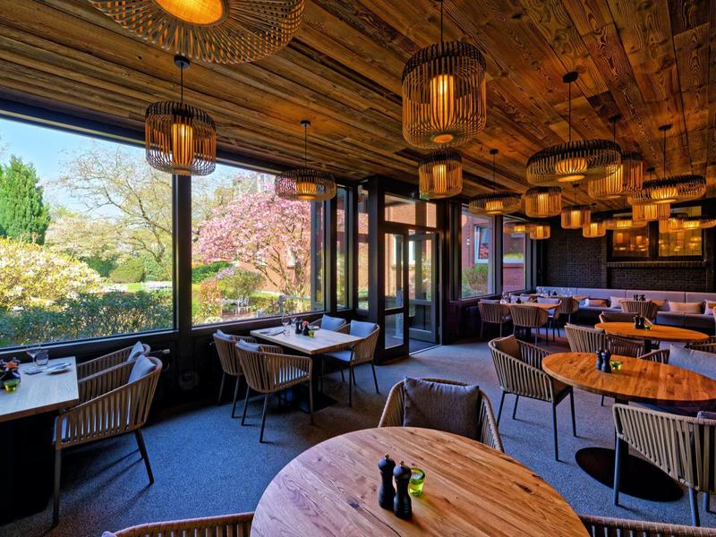 Interno accogliente del ristorante con tavoli in legno e grandi finestre che si affacciano su un giardino.