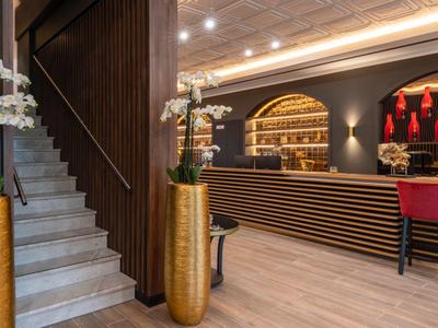 Moderne Hotelbar mit dunkler Theke, roten Stühlen, hellem Holzboden und weißen Orchideen in goldenen Vasen.