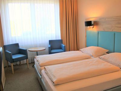 Hell eingerichtetes Hotelzimmer mit zwei Einzelbetten, zwei blauen Sesseln und einem kleinen Tisch.