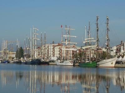 Segelschiffe und Gebäude spiegeln sich am ruhigen Hafenwasser unter klarem Himmel.