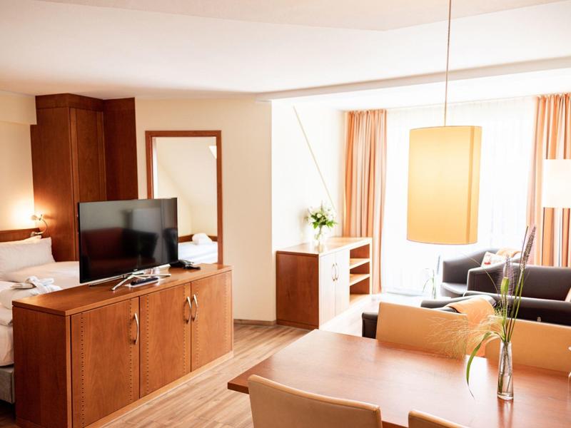 Suite d'hôtel lumineuse avec coin salon, bureau, TV et grande fenêtre avec rideaux.