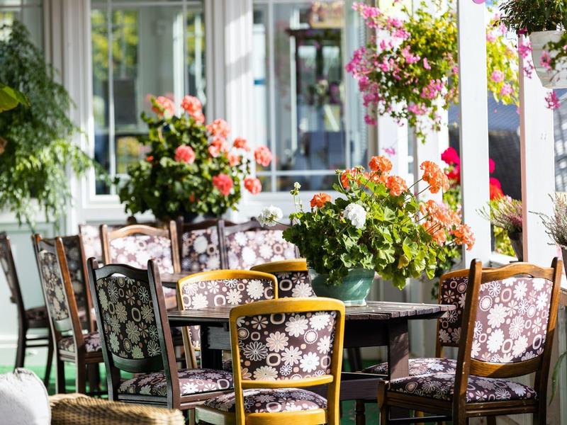 Gemütliche Terrasse mit bunten Blumen, Holztischen und Stühlen mit gemustertem Polster.