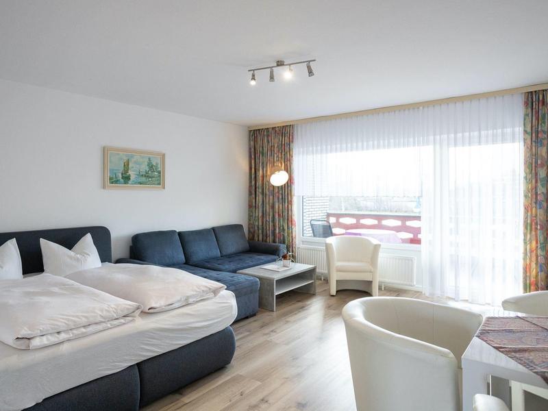 Helles Zimmer mit Doppelbett, blauer Couch, weißen Sesseln und großem Fenster mit Gardinen.