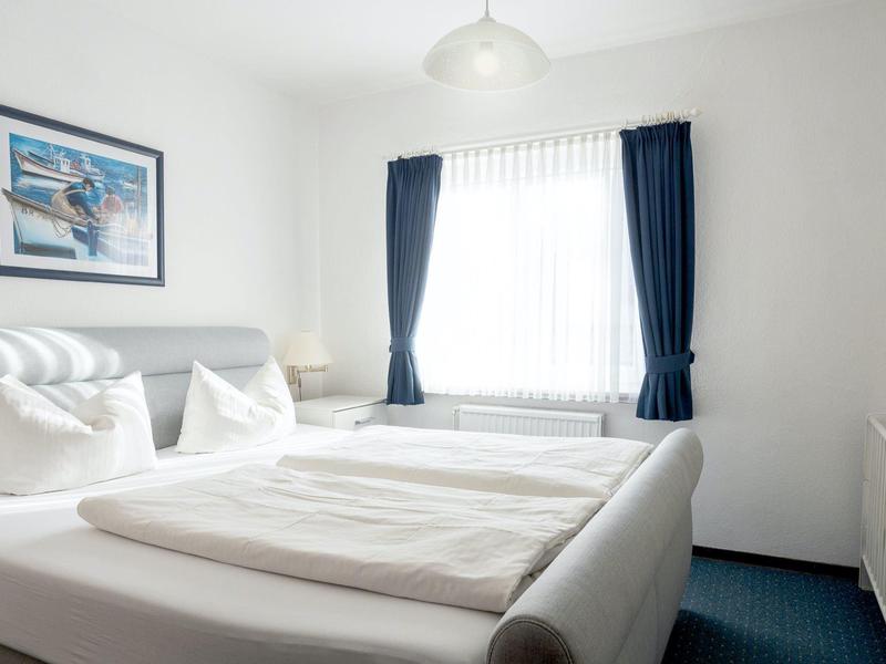 Helles Schlafzimmer mit weißem Bett, blauen Vorhängen und einem maritimen Bild an der Wand.